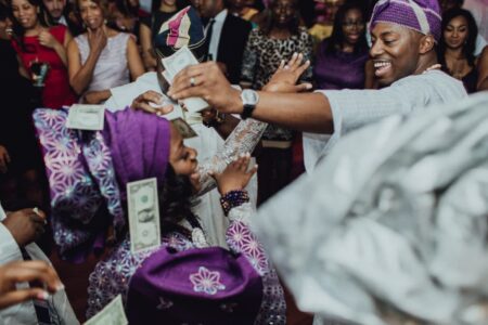 Nigerians in vibrant purple attire dance and smile at a party, joyfully celebrating as money is tossed in the air.