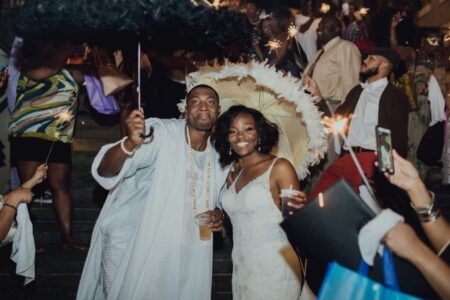 A smiling couple in wedding attire stands under decorated umbrellas, holding drinks amid sparklers at a festive celebration.