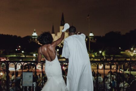 Bride and groom in white wave from a balcony at night, with a lit cathedral and city lights behind them.