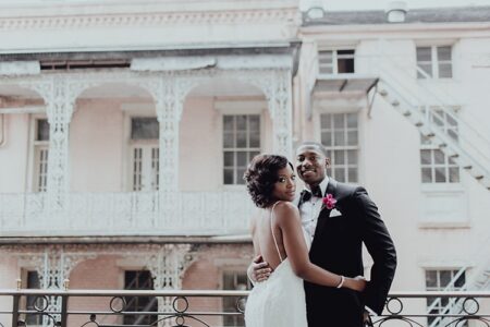 Bride and groom embrace and smile on a balcony with ornate ironwork, pale pink building in the background.