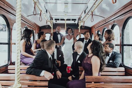 Bride and groom smile on a trolley with their wedding party in black tuxedos and purple dresses, laughing together.