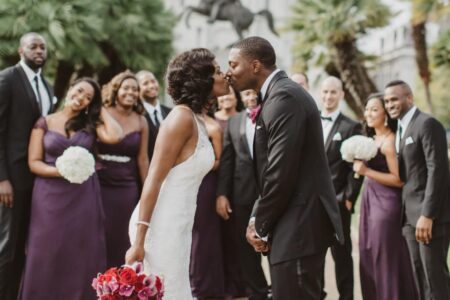 Bride and groom kiss outdoors with smiling wedding party in black suits, purple dresses, holding white bouquets and red roses.