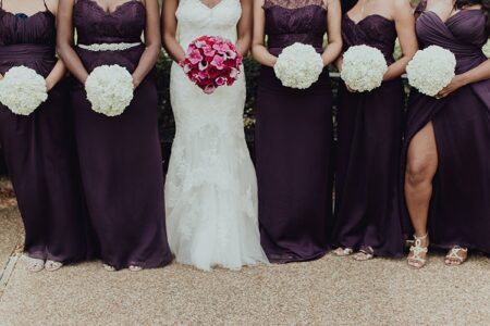 Bridal party lineup: five in dark purple gowns with white bouquets, bride in white lace dress holding pink flowers, shoulders down.