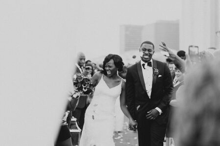 Smiling bride and groom walk hand in hand down the aisle as guests cheer; black and white photo.