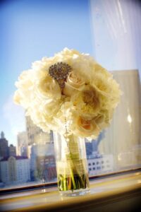 White rose bouquet with jeweled brooch in a glass vase on a windowsill, cityscape and blue sky behind.