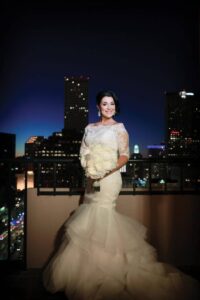 Bride in white gown holding bouquet on a city rooftop at night, with tall buildings and city lights behind her.