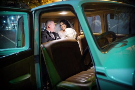 Bride in a white gown and groom in a tuxedo smile together in a vintage car at night, softly illuminated.