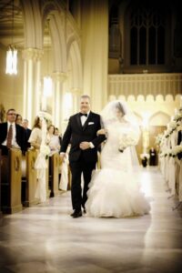 Bride in white gown and veil walks down church aisle with man in tuxedo, surrounded by guests and flowers.