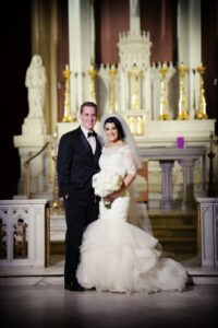 Smiling bride and groom pose before an ornate church altar. Bride holds white flower bouquet, both in classic wedding attire.