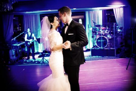 Bride and groom share first dance at wedding reception, smiling as a live band plays on a lit stage in the background.