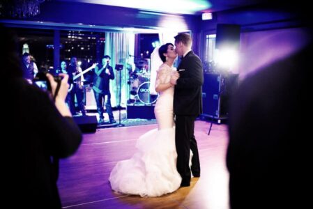 Bride and groom share their first dance as guests watch and a live band plays in an elegant, dimly lit wedding venue.