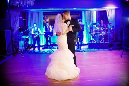 Bride and groom share their first dance in a blue-lit ballroom as a live band plays in the background.