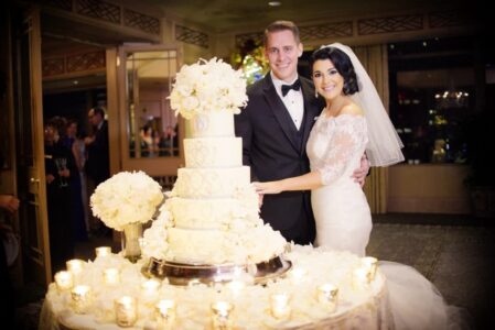 Bride and groom in formal attire smile next to a white floral wedding cake, candles, and flowers in a warmly lit venue.