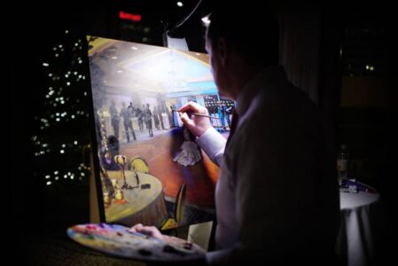 An artist paints a lively formal event on canvas, holding a palette, with tables and people shown under dim lighting.