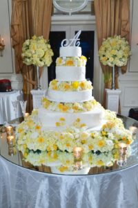 Tall white wedding cake with yellow and white flowers on a mirrored table, surrounded by candles and floral decor.