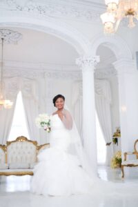 Bride in white gown smiles with bouquet in elegant, ornate room featuring columns, chandeliers, and vintage furniture.