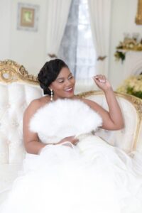 Smiling woman in a white gown sits on an ornate cream sofa with a white fan, gold decor and white curtains behind her.