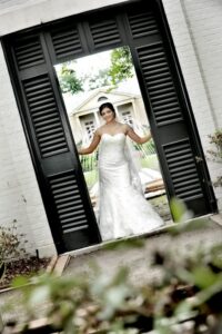 Bride in white dress poses in a large doorway with black shutters, outside a white building surrounded by greenery.