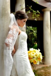 Bride in white lace dress and veil stands by stone columns, holding yellow and white roses, looking down thoughtfully.