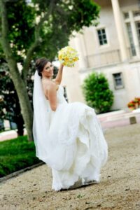 Bride in white dress holding yellow bouquet smiles over her shoulder outside a large building.