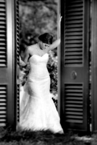 Bride in strapless gown stands between tall open shutters, looking down gently. Black and white photo with soft background.