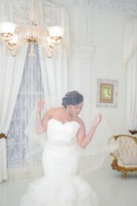 Bride in strapless white gown holds veil, posing indoors amid elegant décor with white curtains, molding, chandelier, and vintage furniture.