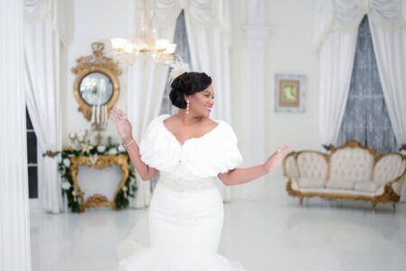 Bride in white gown smiles and poses in an elegant room with gold accents, vintage furniture, and draped curtains.