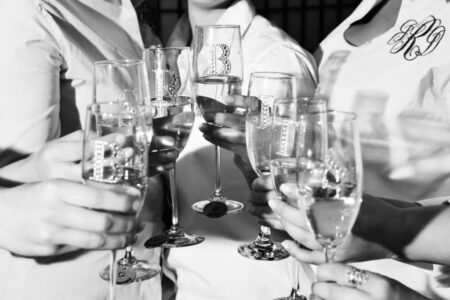 People in light shirts clink champagne glasses with rhinestone initials; black and white photo.