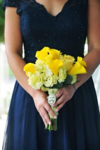 Woman in a navy blue dress holding a bouquet of yellow calla lilies and hydrangeas with jeweled brooch on the stem.