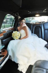 Bride in white dress and veil sits in limousine backseat, holding a yellow and white rose bouquet.