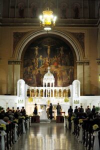 Wedding ceremony in a grand church with high ceilings, ornate decor, large mural, guests seated, and wedding party at the altar.
