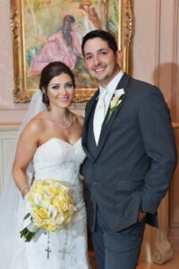 Bride in white dress with yellow-white bouquet stands by groom in gray suit, both smiling indoors with painting behind them.