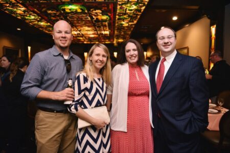 Four adults, two men and two women, smile at an indoor event with stained glass ceilings and dim lighting.