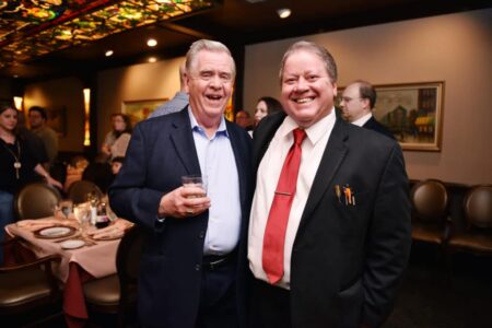 Two men in suits smile at a formal restaurant event; one holds a drink, with people and dinner tables in the background.
