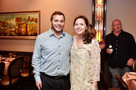 Smiling man and woman in a restaurant, with another man holding a beer. Stained glass window and painting on the wall.