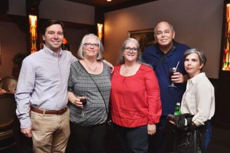 Five adults indoors at a social event, smiling at the camera. Three women in the center, two men on the sides, some holding drinks.