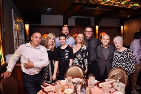 Eight people in semi-formal wear smile together at a restaurant table with drinks, plates, and stained glass in warm lighting.