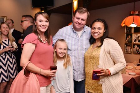 Four people—three adults and a child—smile indoors at a party. Two women hold drinks; food and guests are in the background.