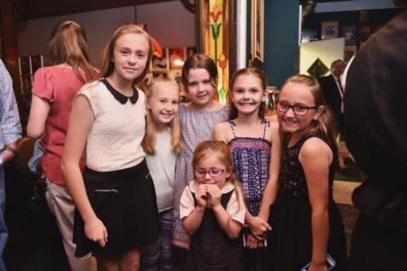 Six young girls smiling and posing together at an indoor event, with people and colorful decor in the background.