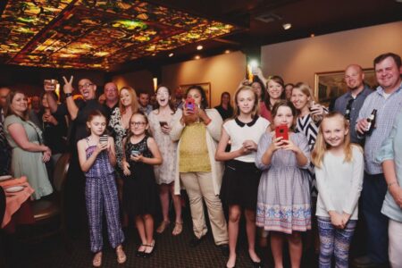 Smiling adults and children stand close indoors, holding phones or drinks in a joyful, celebratory setting.