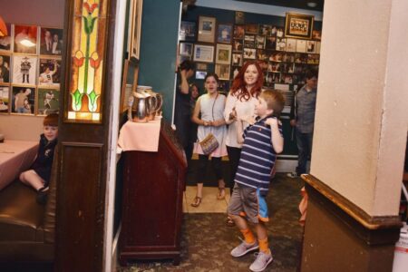 Smiling adults and children gather in a cozy restaurant with framed photos; one boy sits at a booth while others enter from a hallway.