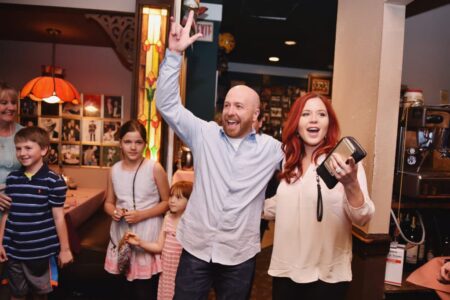 Smiling man and woman wave at an indoor celebration with children, under warm lighting.