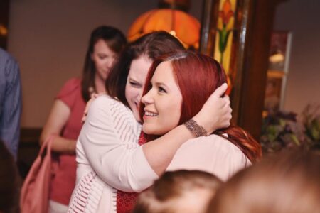 Two women, one with red hair and one with brown hair, smile as they hug indoors with others and warm lighting in the background.