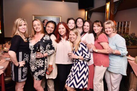 Ten women smile together at a restaurant table, some with drinks, all in stylish outfits, celebrating.