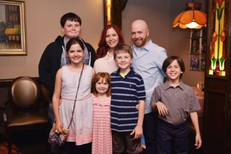 A family of seven poses indoors in warm light, with stained glass and vintage decor visible in the background.