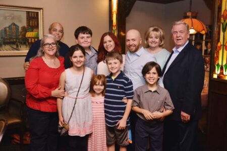 Eleven adults and children smile together in a warmly lit room, suggesting a cheerful family gathering.
