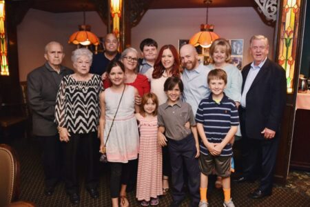Thirteen people of various ages smile together in a warmly lit room with stained glass lamps and ornate wood decor.