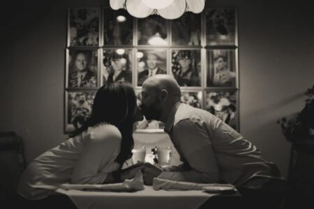 A couple kisses at a dimly lit restaurant, with black and white photos on the wall creating a nostalgic atmosphere.