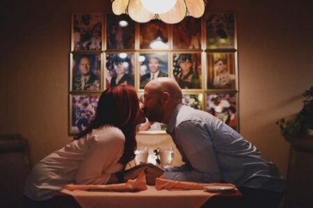 Couple leans in for a kiss at a dining table, with military photo frames behind them and warm, intimate lighting.