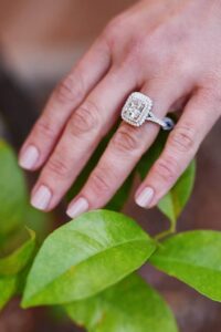 Hand with light pink nails rests on green leaves, displaying a diamond ring with a large center stone and double halo.